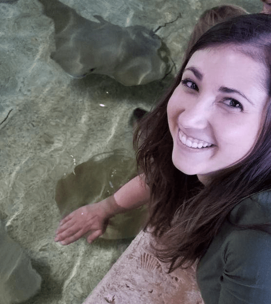 A picture of Nicole Campbell at the aquarium Touch Pool. She has her hand in the water and there is a stingray beneath her hand. She is looking up and smiling at the camera. 