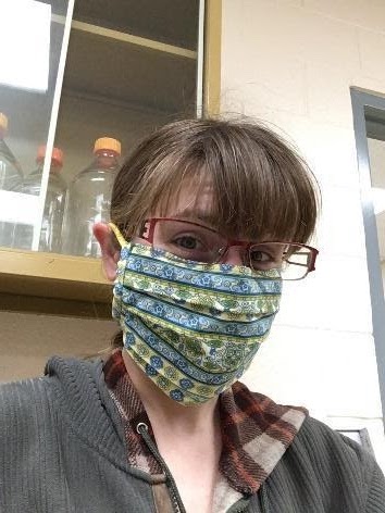 Kara wears a face mask and smiles at the camera. She is in a laboratory. 