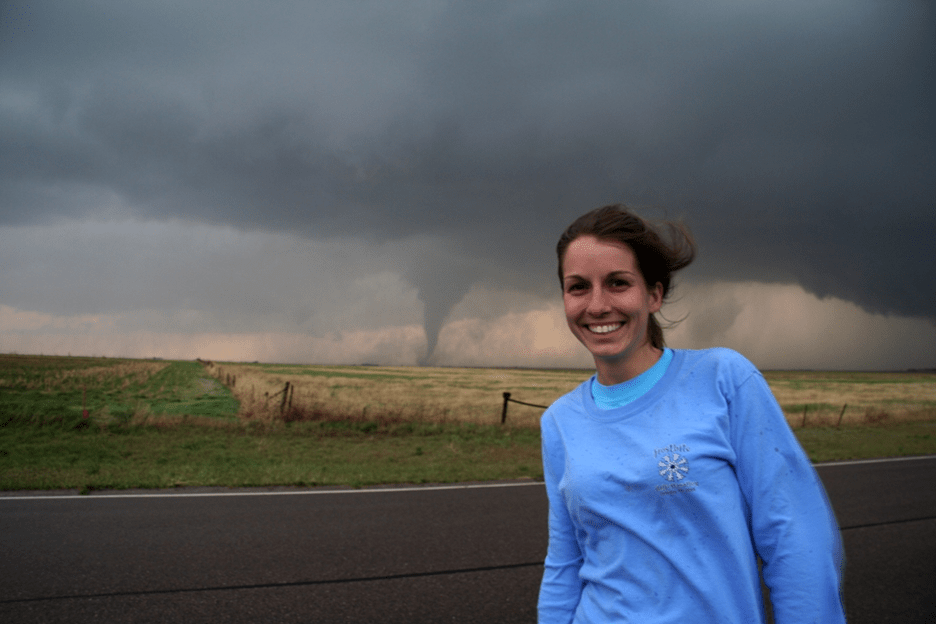 Dr. Kelsey Ellis storm chasing in 2006. Dr. Ellis smiles at the camera, wind blowing in her hair. There is an active tornado in the background. 