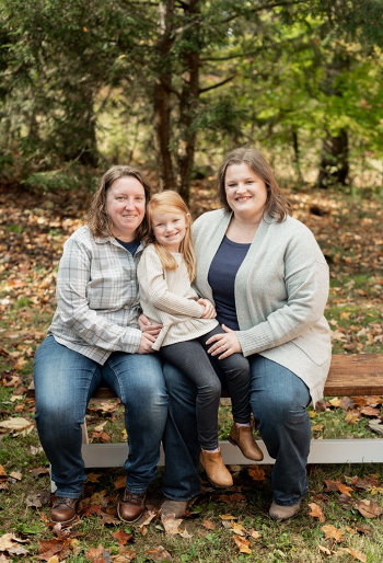 Lesley with her wife sit on a bench. Their and daughter sits between them.