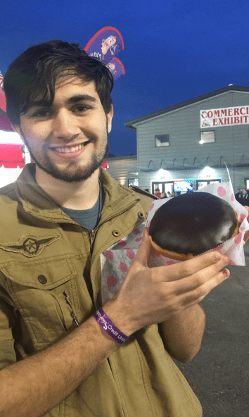 Derrick smiles at the camera. He has brown hair and a thin beard. He is holding up a donut. 