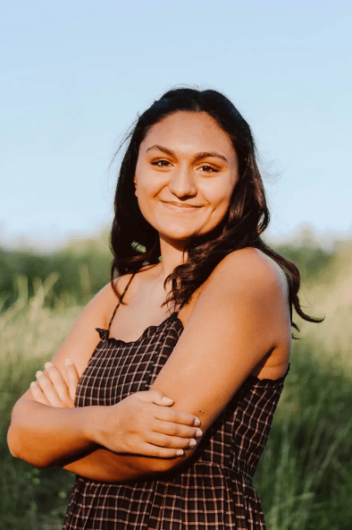 Sydney smiles at the camera. She has black hair and is wearing a black dress with yellow cross-crosses patterned on the front..