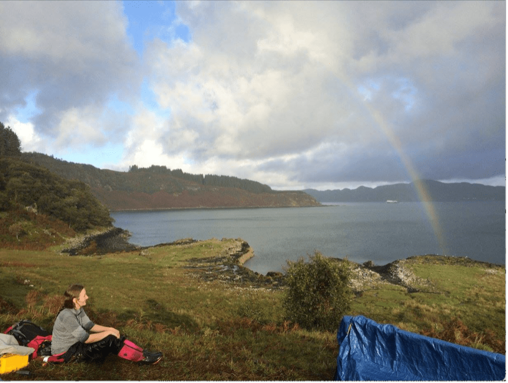 Dr. Findlay is sitting on an island staring out to sea. She is looking for harbor porpoises as part of a scientific survey during a rainstorm. She is soaking wet from being rained on, but a rainbow has formed in the background as the sun has finally started to come out. 