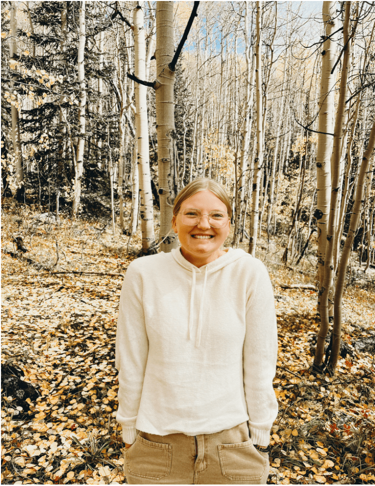A picture of Aspen in the woods.She has light blonde hair and is wearing a while sweater. She stands with her hands in her pockets, smiling at the camera