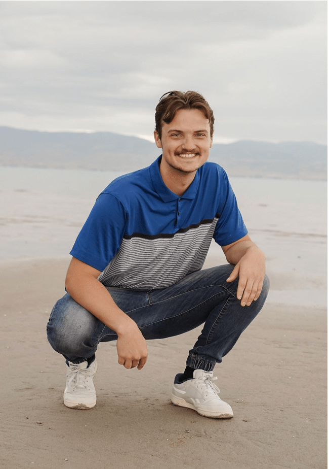 A picture of D.J. crouching on beach sand. He has dark brown hair and a mustache. He is wearing a blue shirt with stripes on the the bottom.