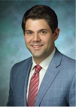 A headshot of Dr. Huhn. He has dark brown hair and is wearing a grey suit and smiling at the camera.