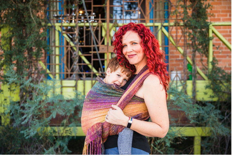 Dr. Lela Rankin is a white woman with red curly hair. She is smiling and wearing her three-and-a-half-year-old son on her front using a hand-woven wrap. Her son is smiling and sticking out his tongue. The background is a brick building with greenery and fencing. The wrap is gold, green, and purple. 