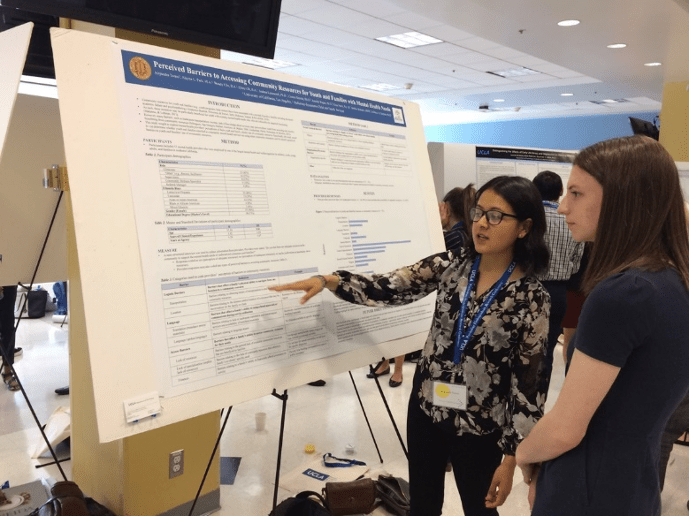 A picture of Alejandra presenting a poster at a conference. She points at the poster while someone stands to listen in front of the poster.