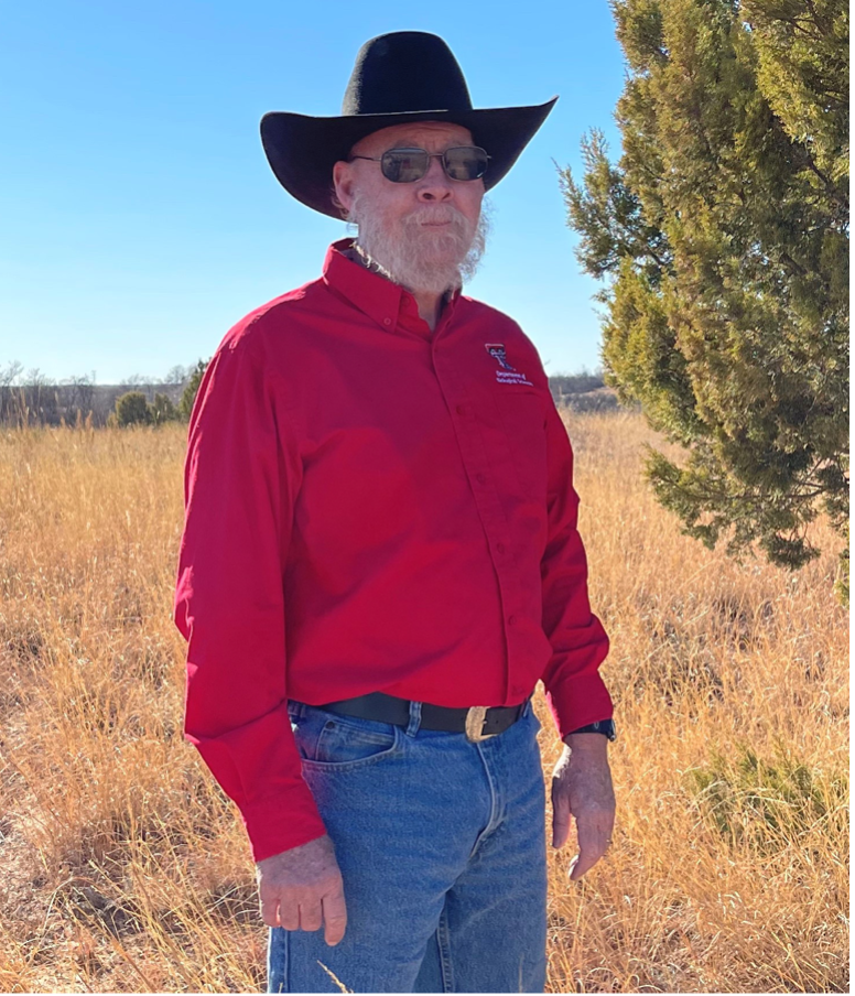 A picture of Dr. Robert Bradley. Dr. Bradley stands in a dry prairie. He wear a cowboy hat and sunglasses. He has a grey beard and mustache. 